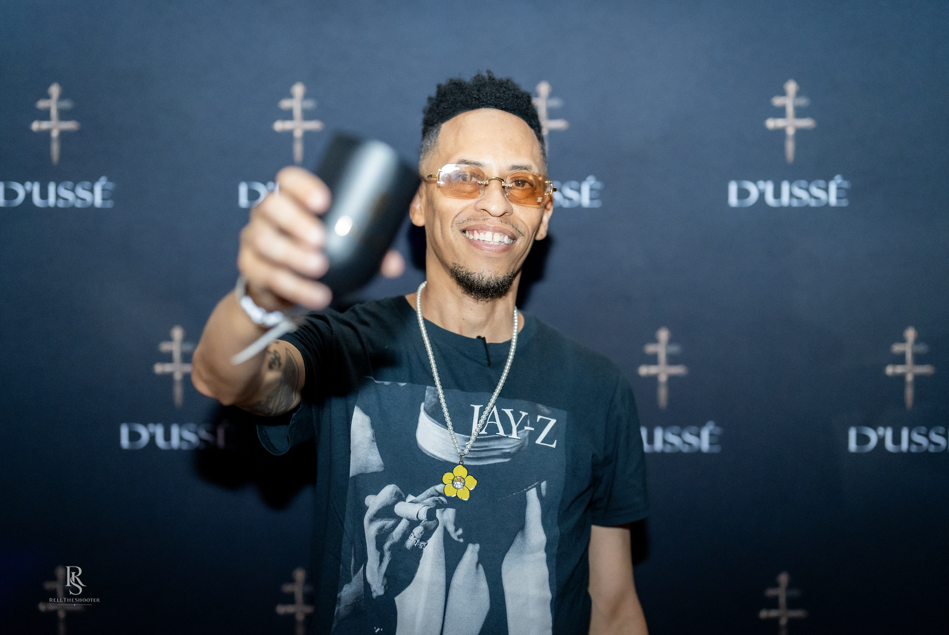 A smiling man wearing a Jay-Z t-shirt and glasses, holding a microphone and posing in front of a D'usse backdrop.