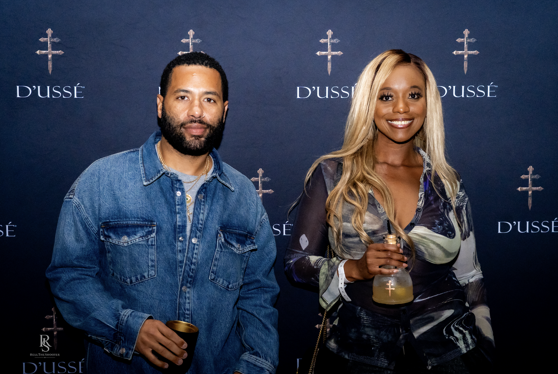 Two people, a man with a beard and a woman with long blonde hair, standing in front of a backdrop with the D'usse logo.