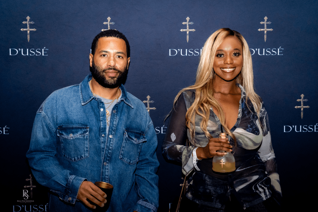 Two people, a man with a beard and a woman with long blonde hair, standing in front of a backdrop with the D'usse logo.