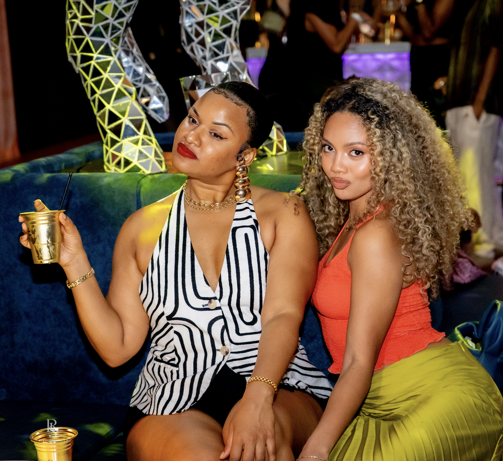 Two women in stylish outfits sitting on a couch in a colorful, modern setting, holding drinks.
