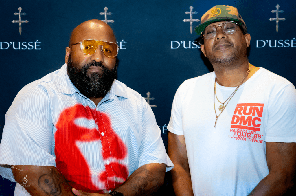 Two men wearing sunglasses and hats, one with a "Run DMC" t-shirt, posing in front of a "D'usse" backdrop.