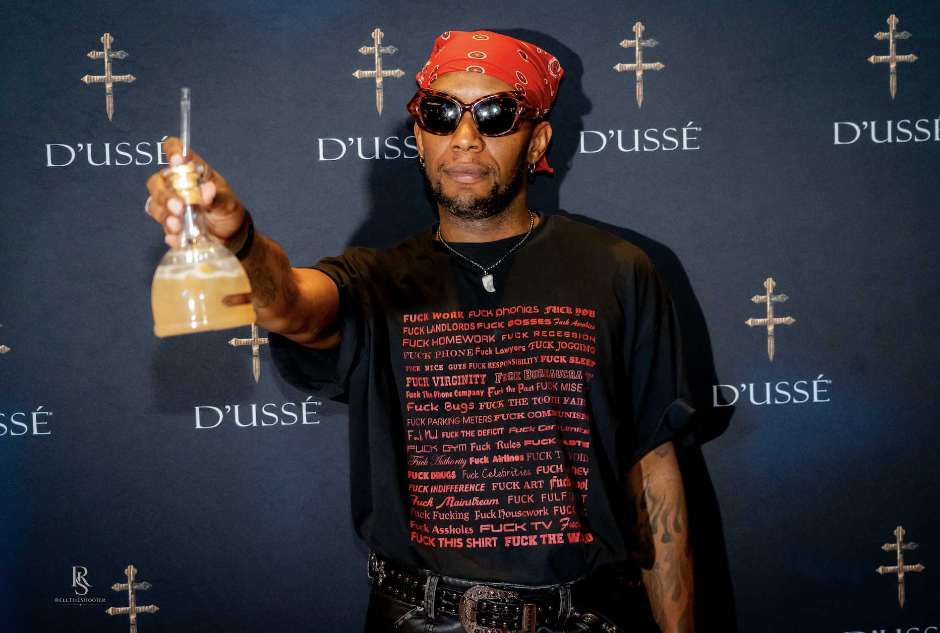 A man wearing a red bandana, sunglasses, and a t-shirt with profanity-laden text, holding a glass and posing in front of D'usse branding.