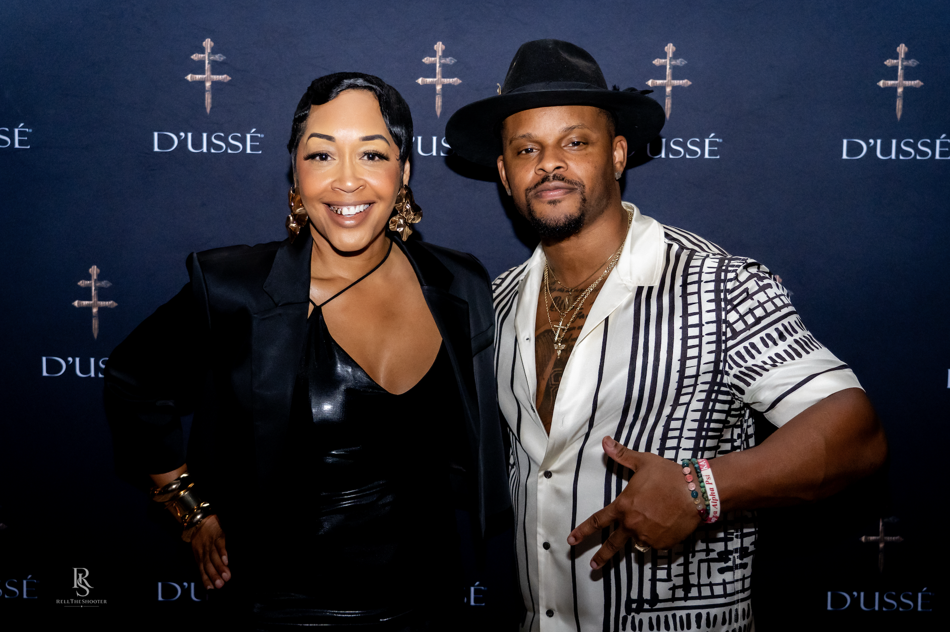 Two well-dressed individuals, a woman in a black outfit and a man in a striped suit, posing in front of a D'usse backdrop.