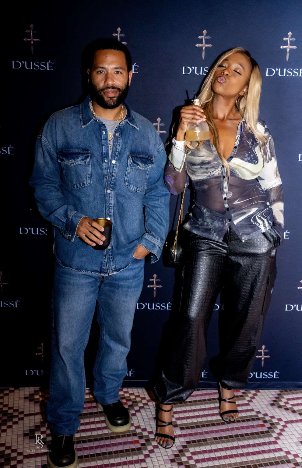 Two people, a man with a beard and a woman with long blonde hair, standing in front of a backdrop with the "D'usse" logo, both wearing denim and casual clothing.