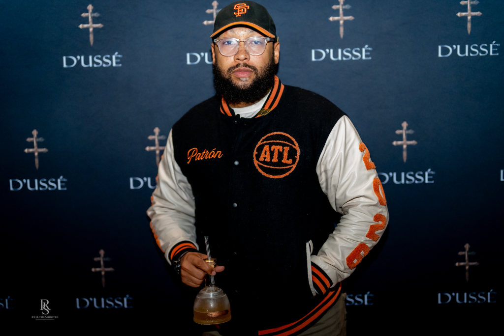 A man wearing a black and orange baseball cap and jacket with the ATL logo, holding a glass, standing in front of a backdrop with the D'usse logo.