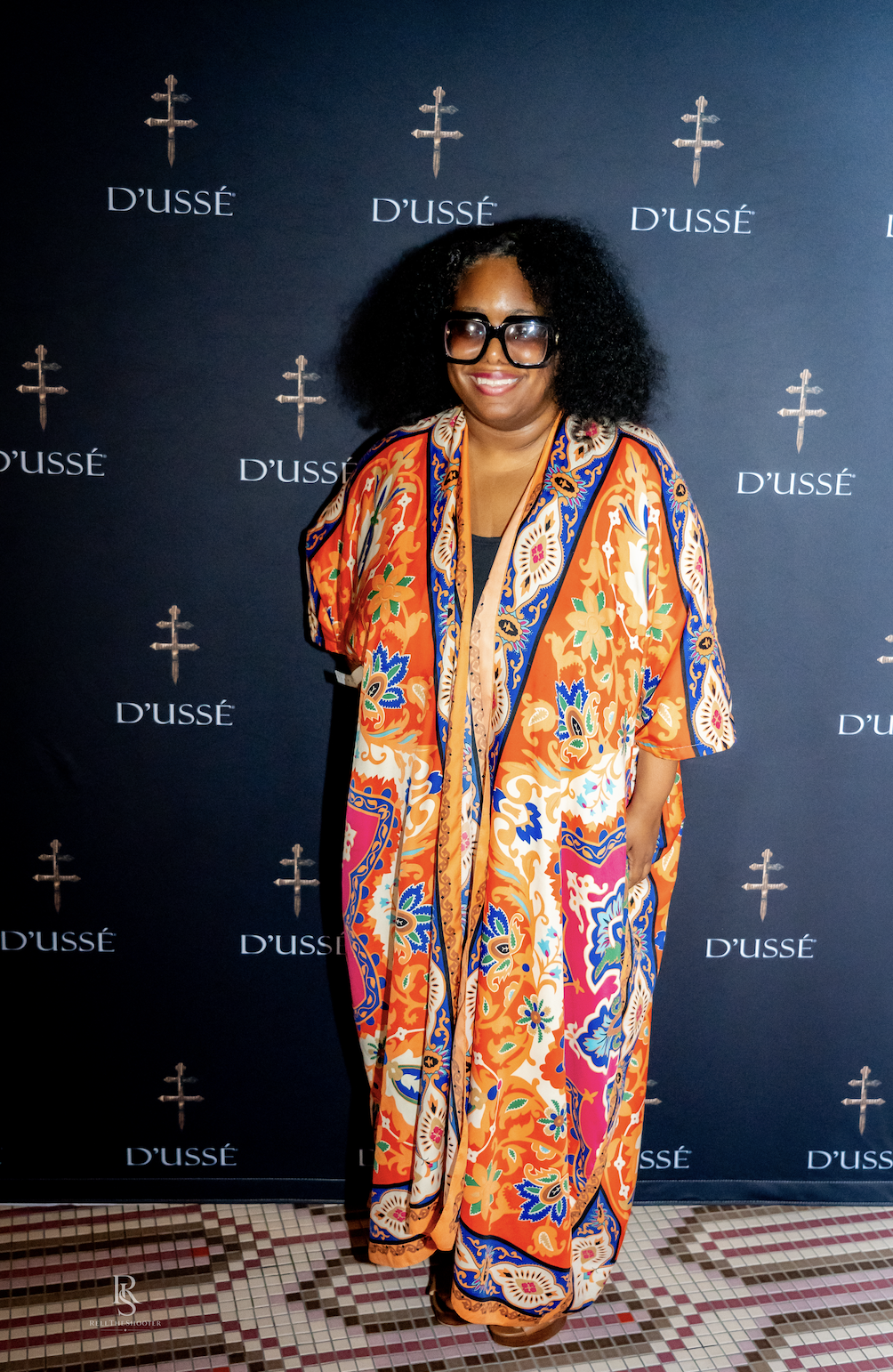 A woman wearing a colorful, patterned robe and large sunglasses stands in front of a backdrop with the D'usse logo.