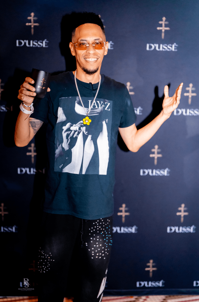 A smiling man wearing a Jay-Z t-shirt and holding a Dussé liquor bottle, standing in front of Dussé branding.