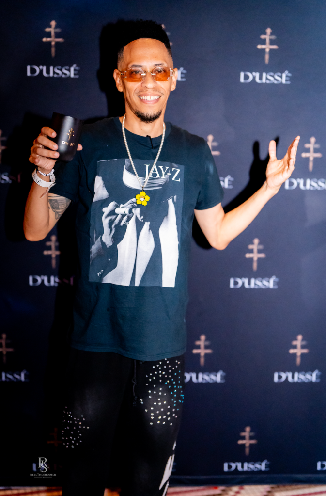 A smiling man wearing a Jay-Z t-shirt and holding a Dussé liquor bottle, standing in front of Dussé branding.
