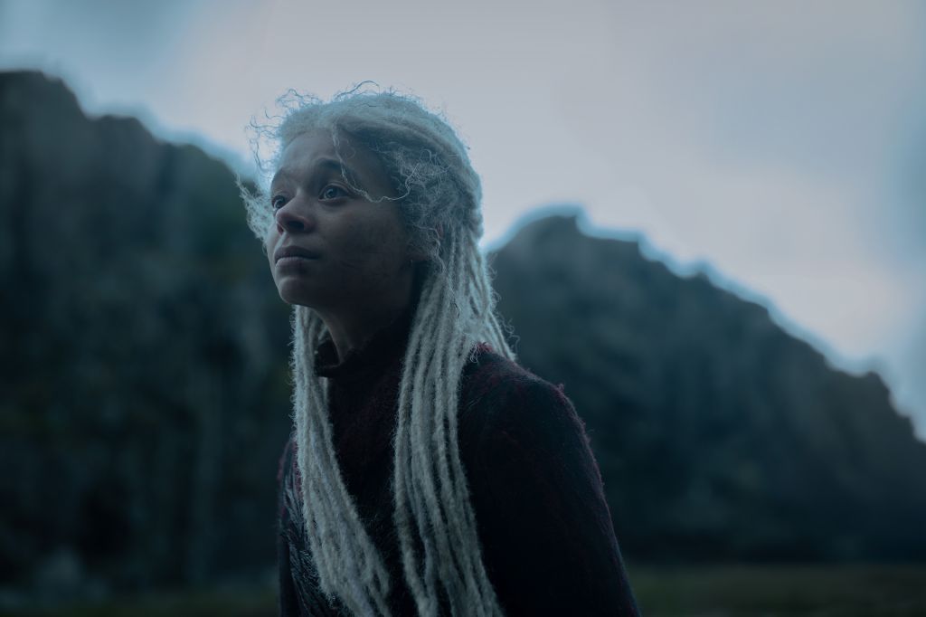 A person with long, wavy gray hair standing in a mountainous, overcast landscape, looking pensive.