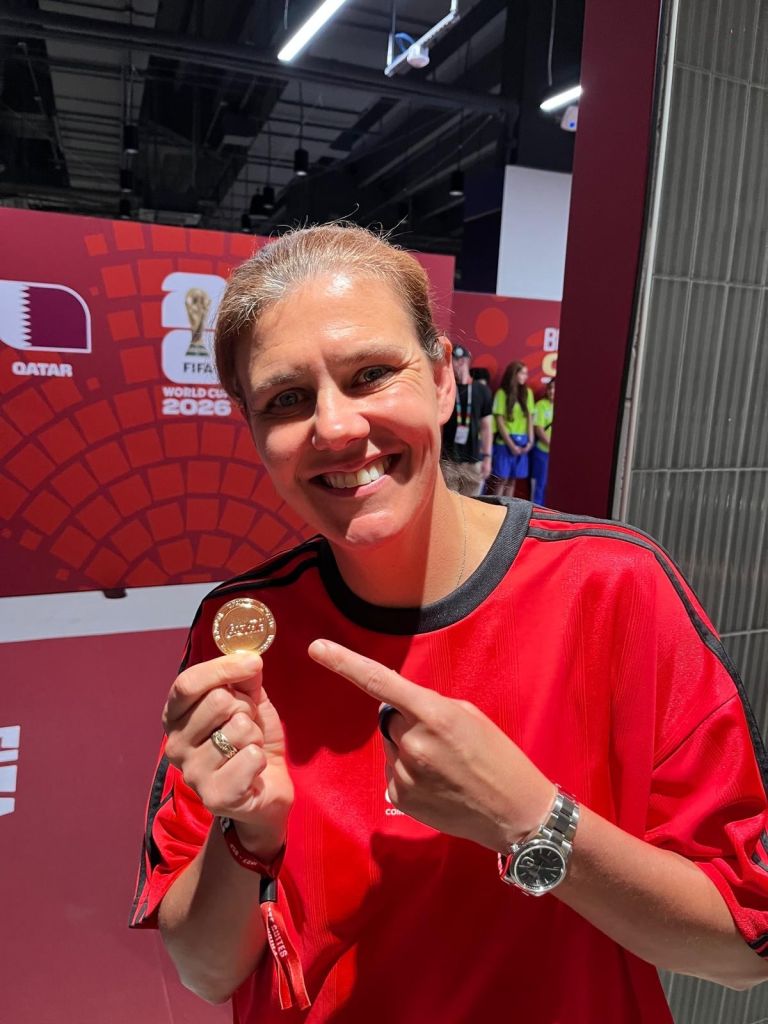 A smiling woman wearing a red jersey and holding a gold medal in front of a FIFA World Cup 2022 backdrop.