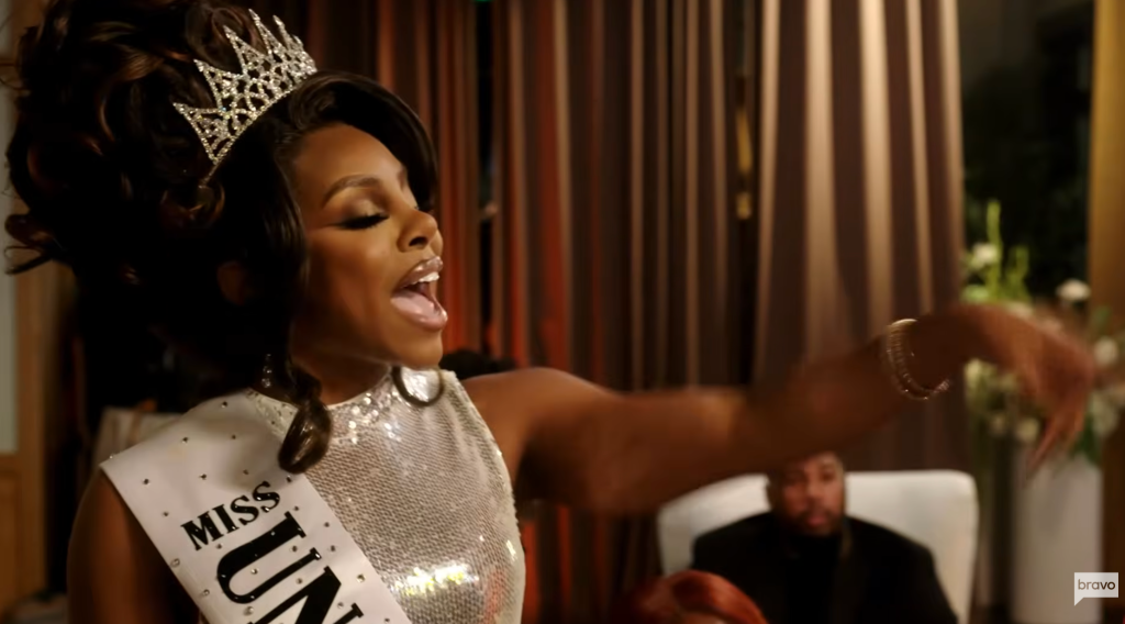 A person wearing a crown and sash labeled "Miss Unity" posing dramatically in a room with curtains.