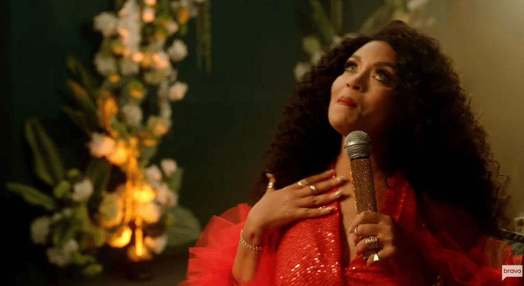 A woman with curly dark hair wearing a red sequined dress sings into a microphone on a stage with floral arrangements.