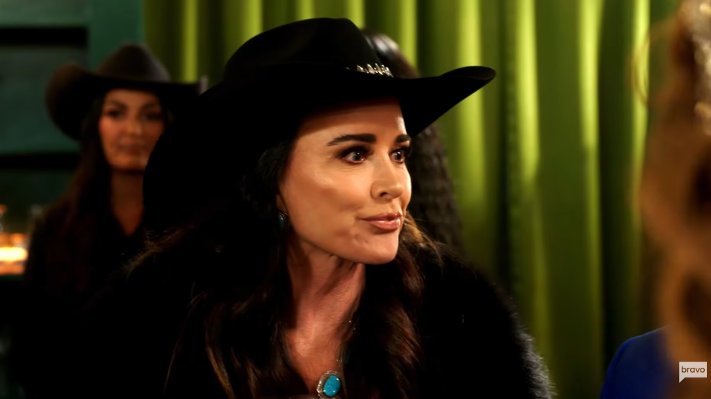 A woman wearing a black cowboy hat and turquoise jewelry stands in a dimly lit room with green curtains in the background.