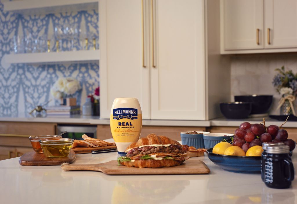 A kitchen counter with various food items, including a Hellmann's Real mayonnaise bottle, a sandwich, grapes, lemons, and other fresh produce.
