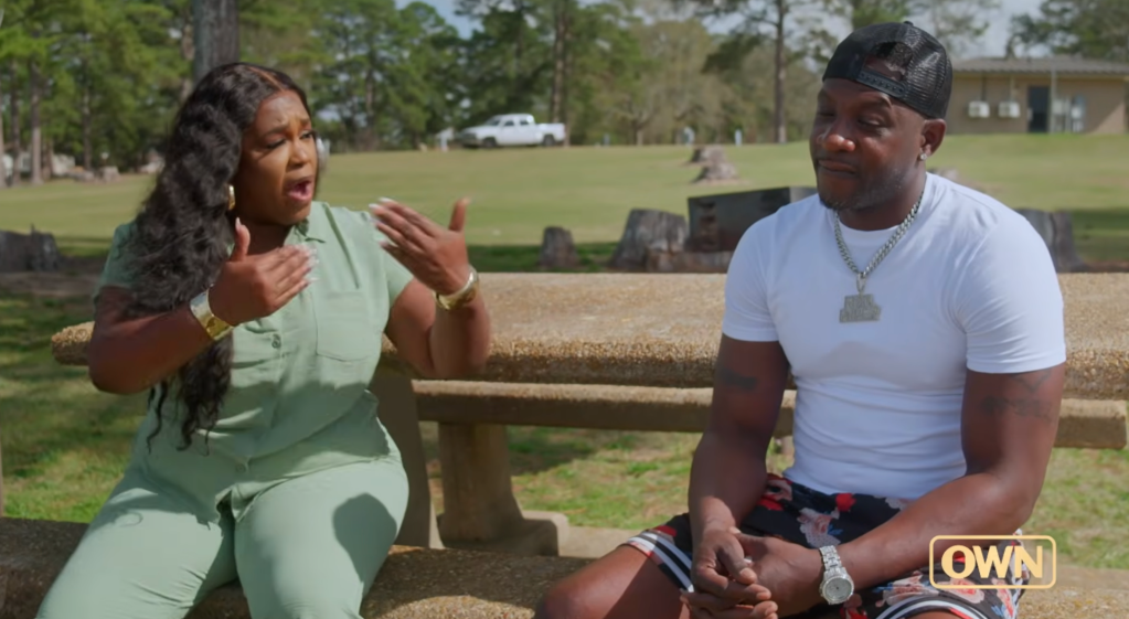 Two people, a woman in a mint green outfit gesturing animatedly and a man in a white shirt and cap, sitting on a bench in a grassy outdoor setting.