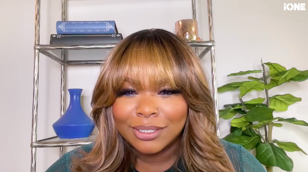 A woman with long, wavy blonde hair and bangs smiling at the camera in front of a shelving unit with decorative items.