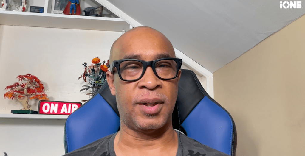 A bald man with glasses wearing a blue gaming chair, speaking into a microphone with an "On Air" sign visible in the background.