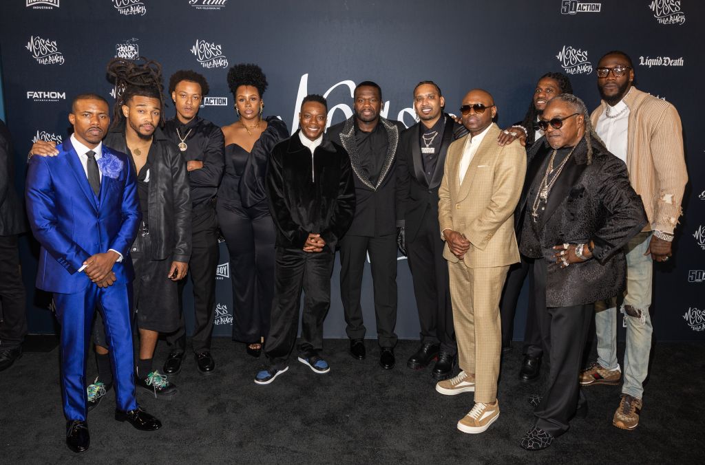 A group of Black men dressed in formal attire posing together at an event, with various logos and branding visible in the background.