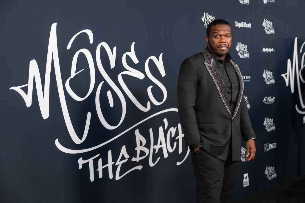 A Black man wearing a black suit and patterned shirt stands in front of a large black and white sign that reads "MOSS THE BLACK".