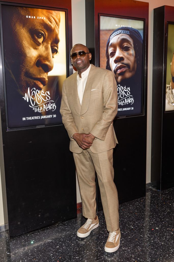 A man in a beige suit standing in front of movie posters for the film "Moss: The Black", which appears to be a film he is promoting.