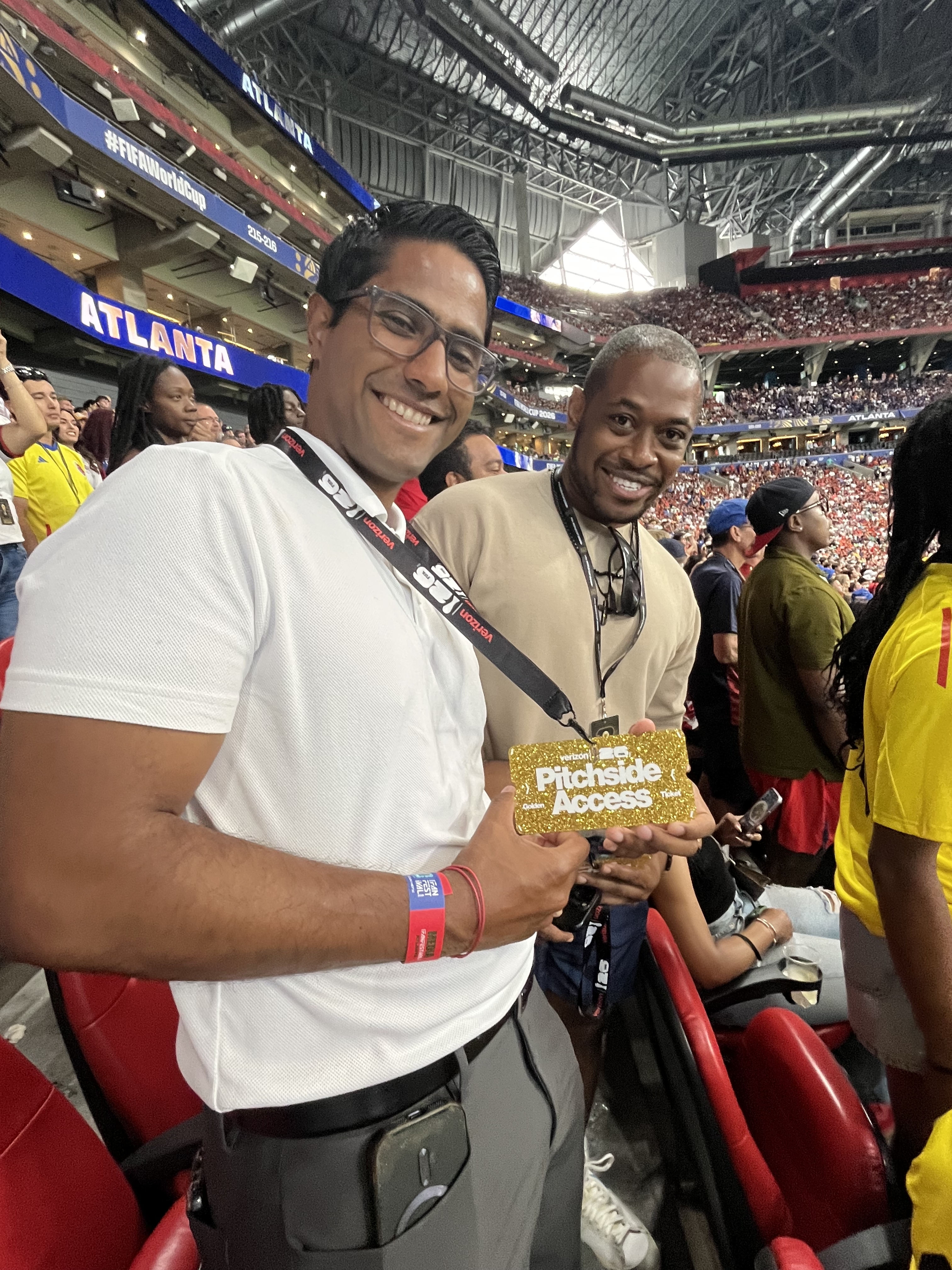 Two men smiling and holding a "Pitchside Access" credential at a crowded sports stadium.