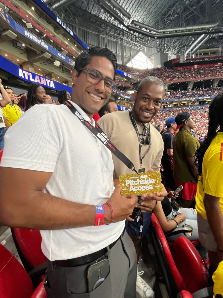 Two men smiling and holding a "Pitchside Access" credential at a crowded sports stadium.