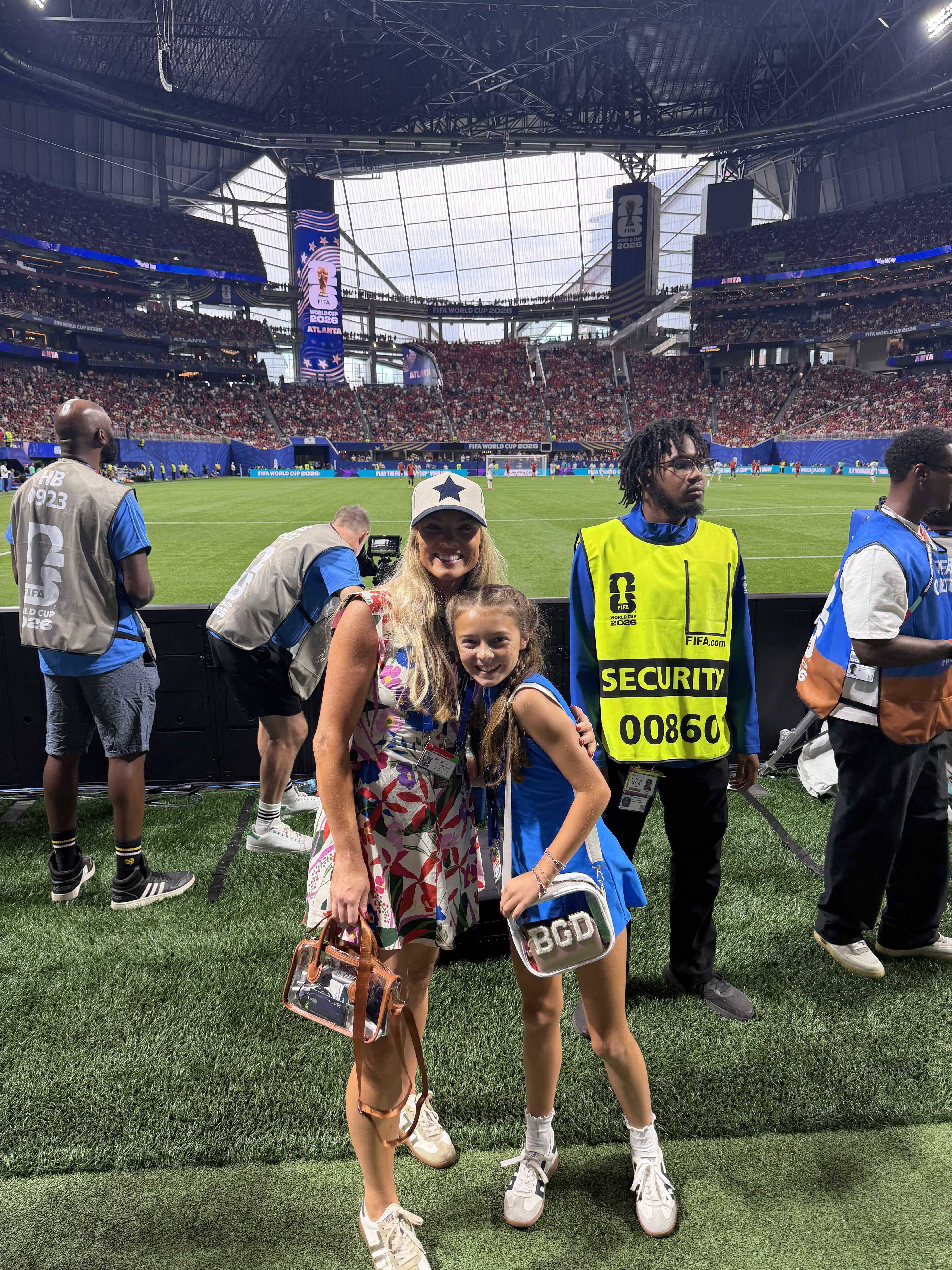 A large indoor stadium filled with spectators. Two women, one wearing a security vest, stand on the field. The stadium displays the FIFA World Cup logo.