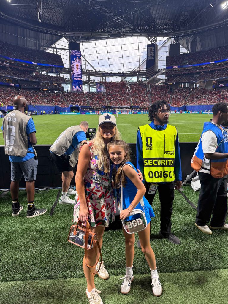 A large indoor stadium filled with spectators. Two women, one wearing a security vest, stand on the field. The stadium displays the FIFA World Cup logo.