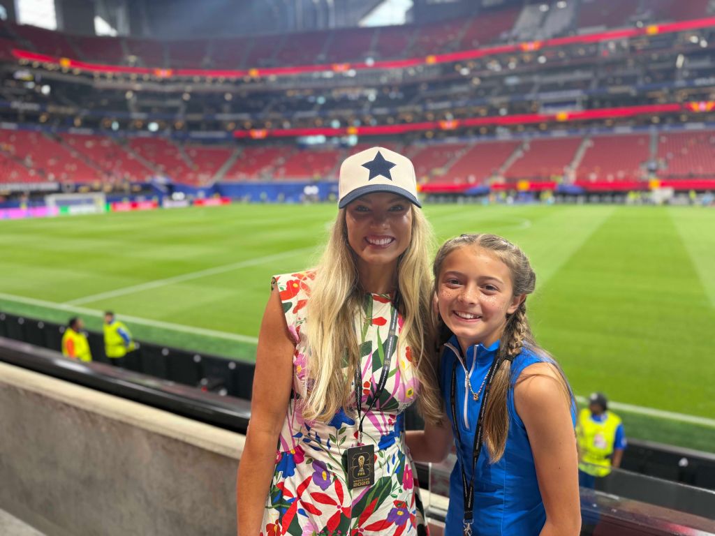 Two young women smiling at a large sports stadium, one wearing a hat with a star logo.