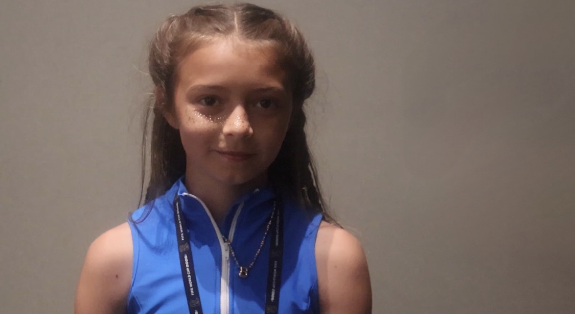 A young girl with long brown hair wearing a blue sleeveless top, looking directly at the camera with a neutral expression.