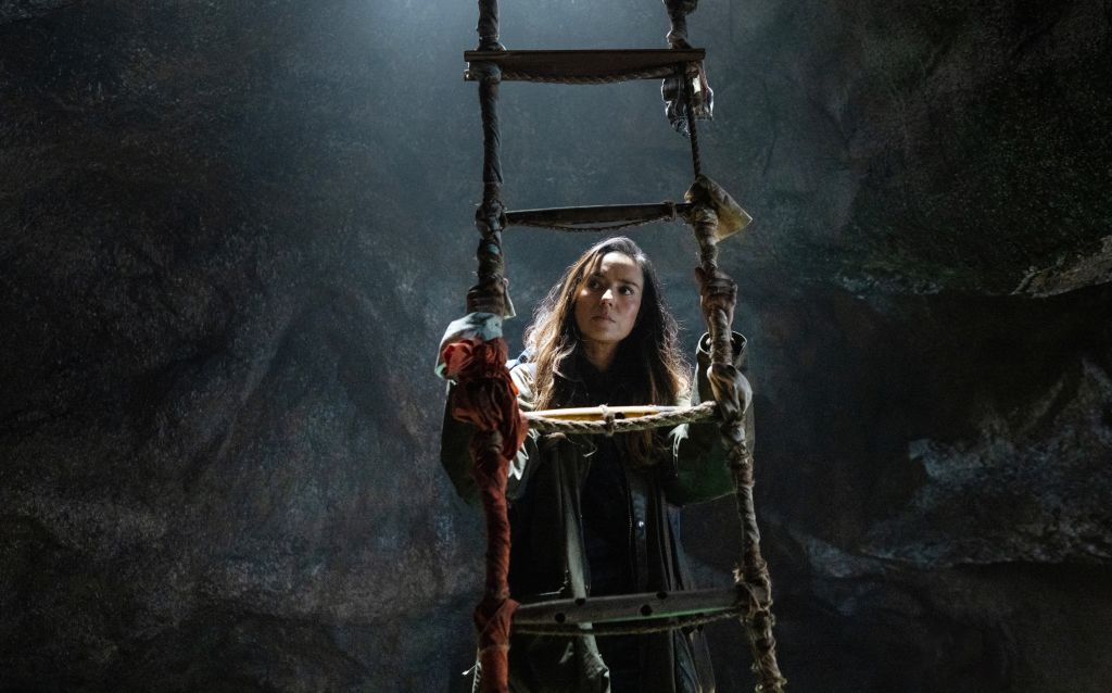A person in dark clothing and a red cloak stands on a rope ladder in a dark, misty cave.