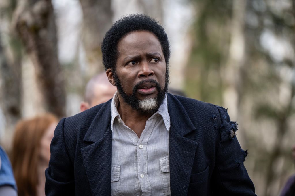 A man with a beard and afro hairstyle wearing a dark jacket and light shirt, standing in a wooded area.