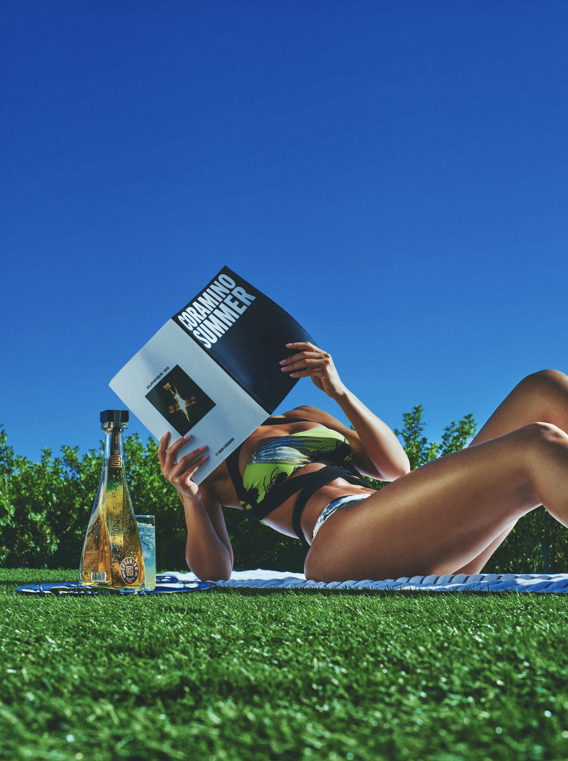 A person reading a book while relaxing on a blanket in a grassy field, with a bottle of alcohol nearby.