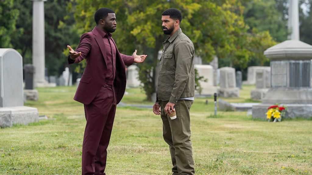 Two men in formal attire conversing in a cemetery setting with gravestones and greenery in the background.