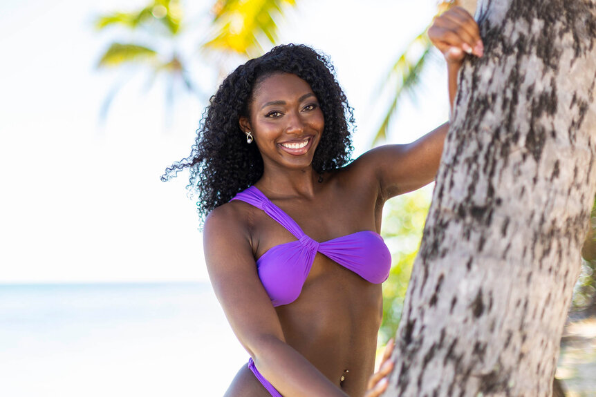 A smiling woman in a purple bikini top leans against a palm tree in a tropical setting.