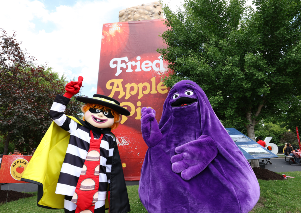 Two costumed characters, one in a striped outfit and hat, the other in a large purple costume, standing in front of a large sign that says "Fried Apple Pie".