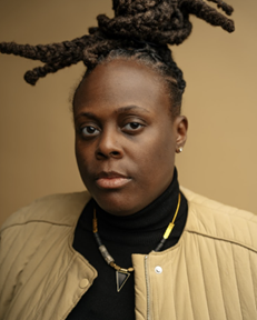 A person with dreadlocked hair wearing a beige jacket and black shirt.