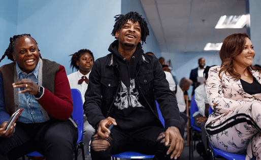 A group of people, including a man with dreadlocks, sitting and smiling in what appears to be a school or office setting.