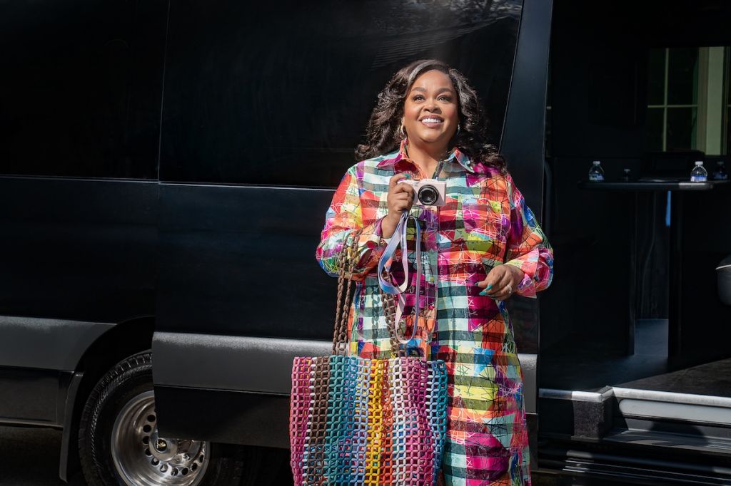 A smiling woman in a colorful plaid coat holding a camera, standing in front of a black vehicle.