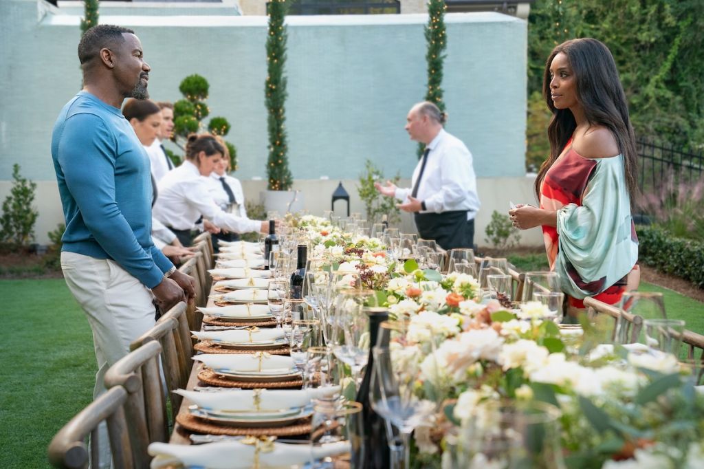 A formal outdoor dinner party with a long table set with white flowers, wine glasses, and place settings. Several people are visible, including a man in a blue shirt and a woman in a red dress.