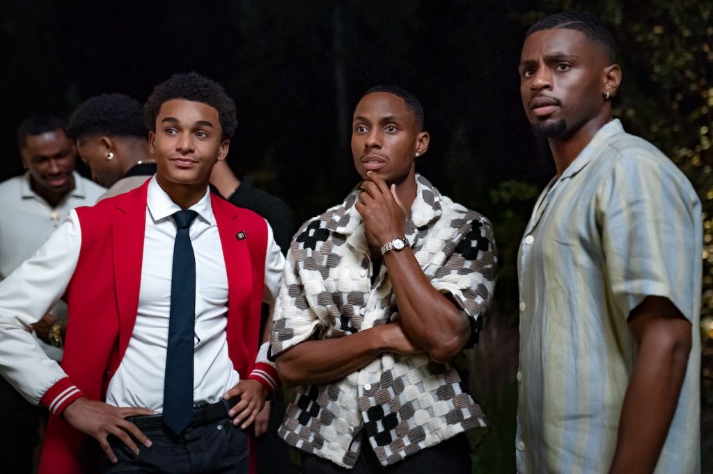 Three young Black men standing together in a dimly lit outdoor setting, one wearing a red jacket and tie, another in a patterned shirt, and the third in a light-colored shirt.