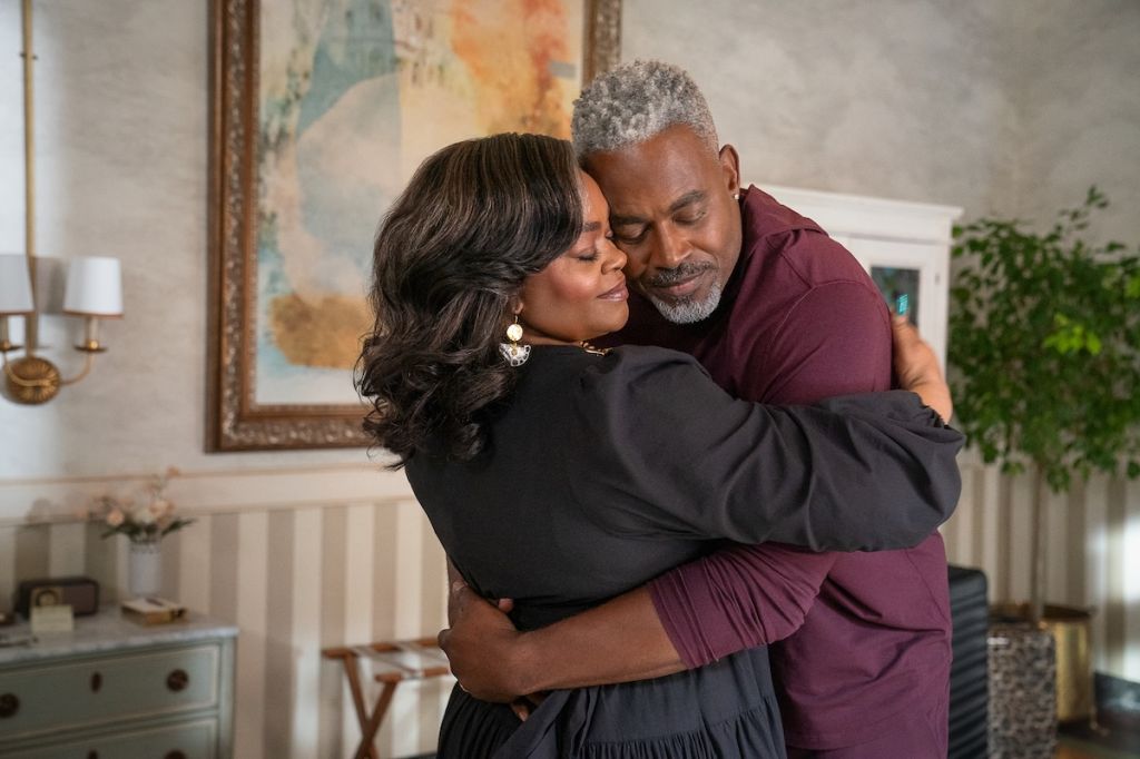 A man and woman embrace tenderly in a cozy living room setting, the woman wearing a black dress and the man wearing a burgundy shirt.
