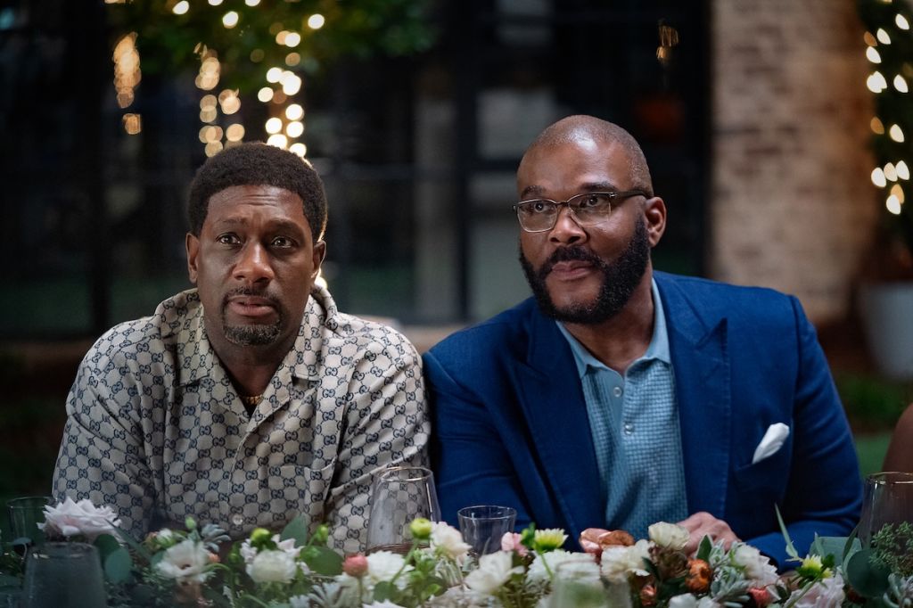Two Black men sitting at a table, one wearing a patterned shirt and the other in a blue suit, surrounded by flowers.