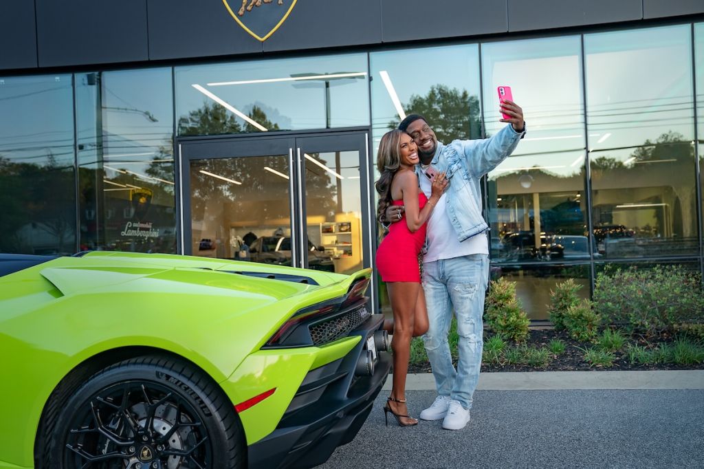 A couple taking a selfie in front of a bright green Lamborghini sports car outside a Lamborghini dealership.