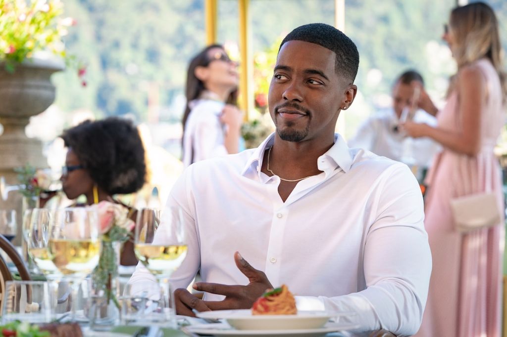 A man in a white shirt sitting at a table in an outdoor setting, with other people visible in the background.