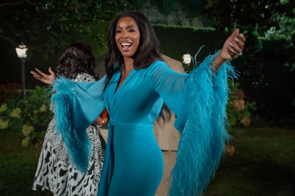 A woman in a teal dress with feathered sleeves smiling and gesturing with her arms outstretched in an outdoor setting.