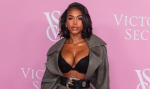 A woman in a black bra and gray coat posing in front of a pink backdrop with "Victoria's Secret" branding.