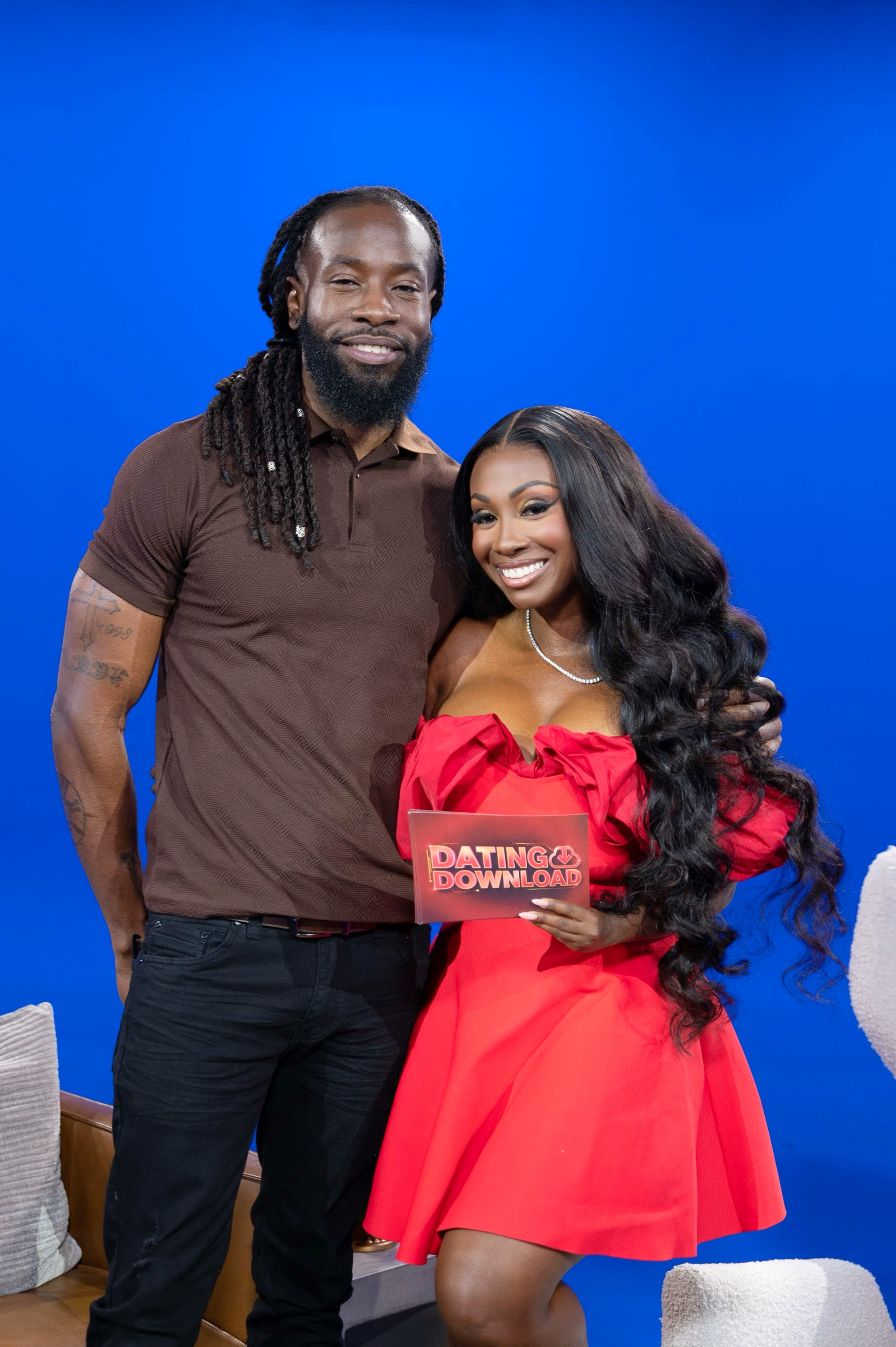 A smiling couple posing together, with the woman holding a "Dating Download" sign in a red dress and the man wearing a brown shirt with dreadlocks.