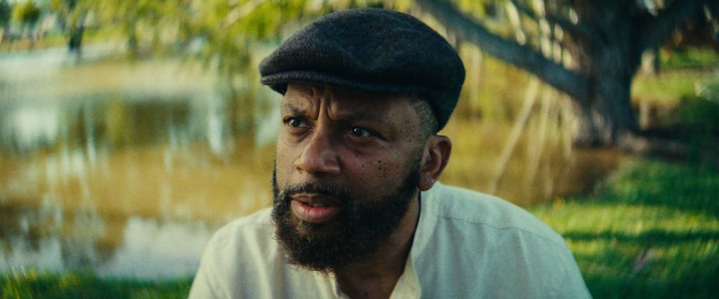 A bearded man wearing a black cap and white shirt, looking directly at the camera with a serious expression, in a natural outdoor setting.