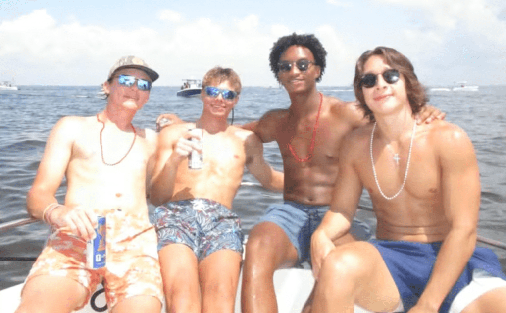 Four people sitting on a boat in the ocean, wearing sunglasses and swimwear, with a boat in the background.